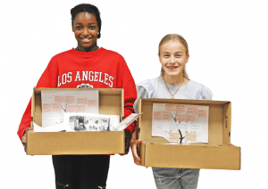 two smiling girls stand holding their family history boxes