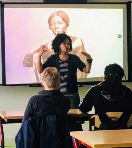 An adult talks to a classroom of students (2 shown) , while standing in front of a photo of Harriet Tubman
