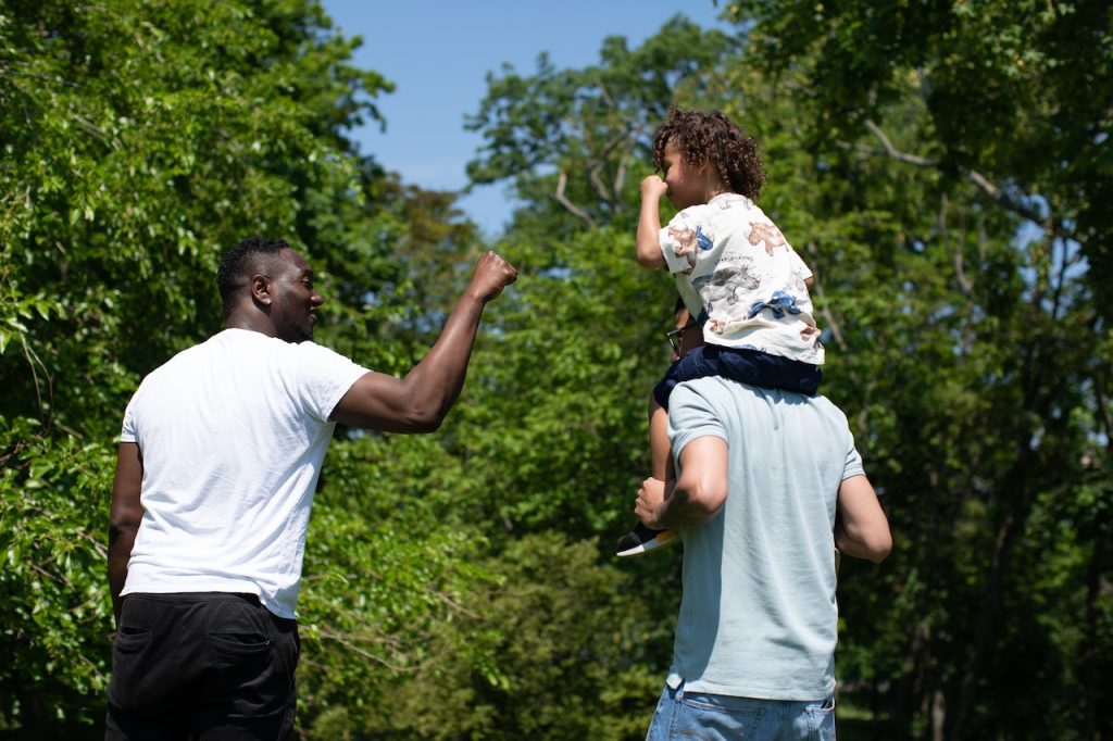 Walking in nature, a child sits on the shoulders of an adult and fist bumps another adult.
